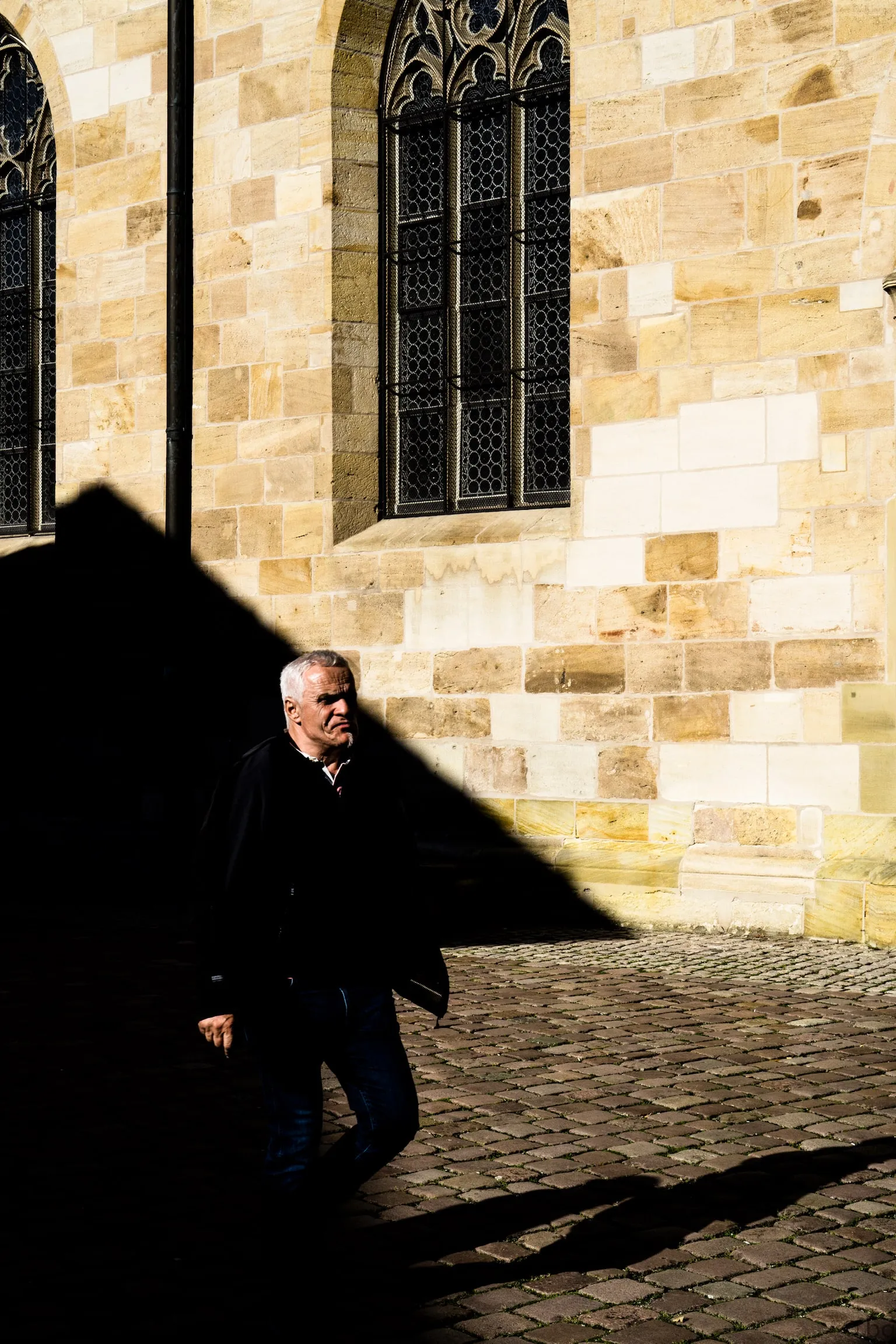 A man walking through deep shadow beside a sunlit stone church wall.