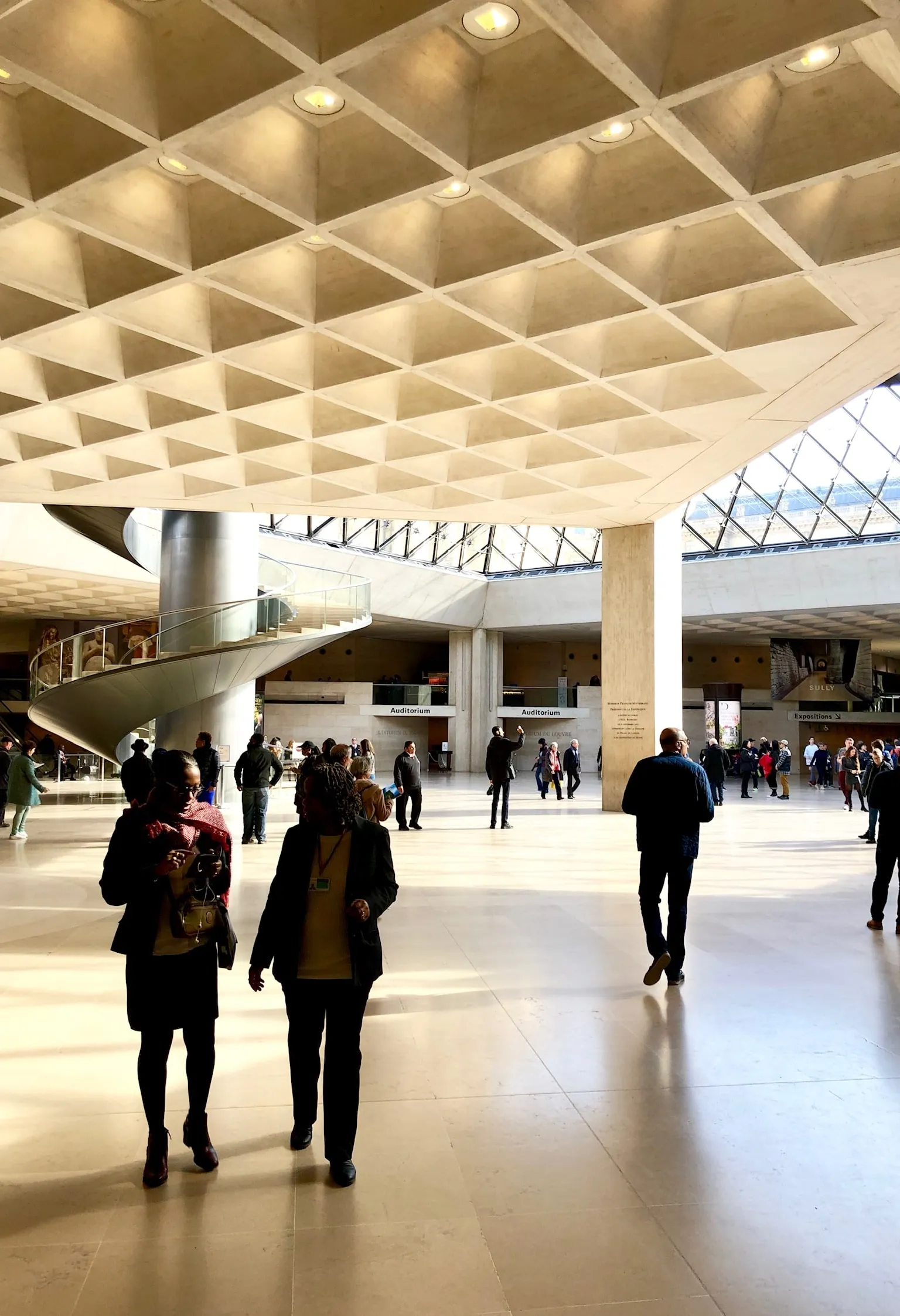 The Louvre concourse: a coffered ceiling above visitors crossing the pale stone floor.