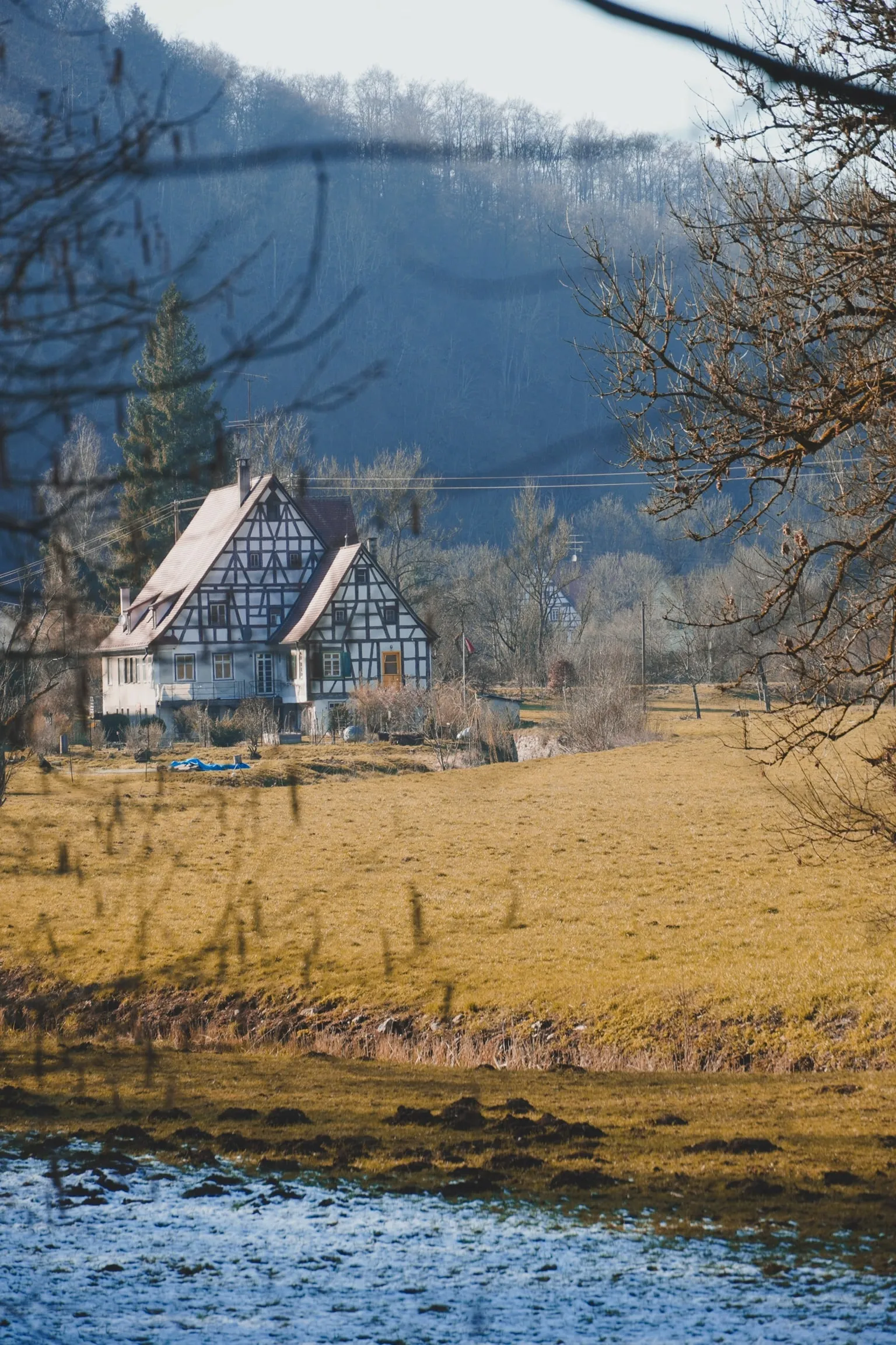 A half-timbered house beside a river, wooded hills rising behind it.